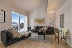 A cozy living room with a black sofa, a grey couch, and a wooden coffee table. There is a large window offering a view of a snowy landscape.