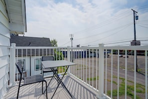 En balkon med et bord og stole, et hvidt gelænder og udsigt til en parkeringsplads samt bygninger i baggrunden.