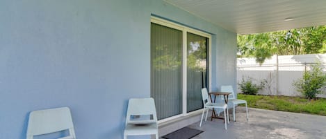 Un espacio de estar al aire libre cubierto con sillas blancas, una mesa, un ventanal corredero y piso de concreto.