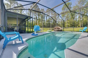 A covered outdoor swimming pool with a view of a forest, a basketball hoop, and lounge chairs.