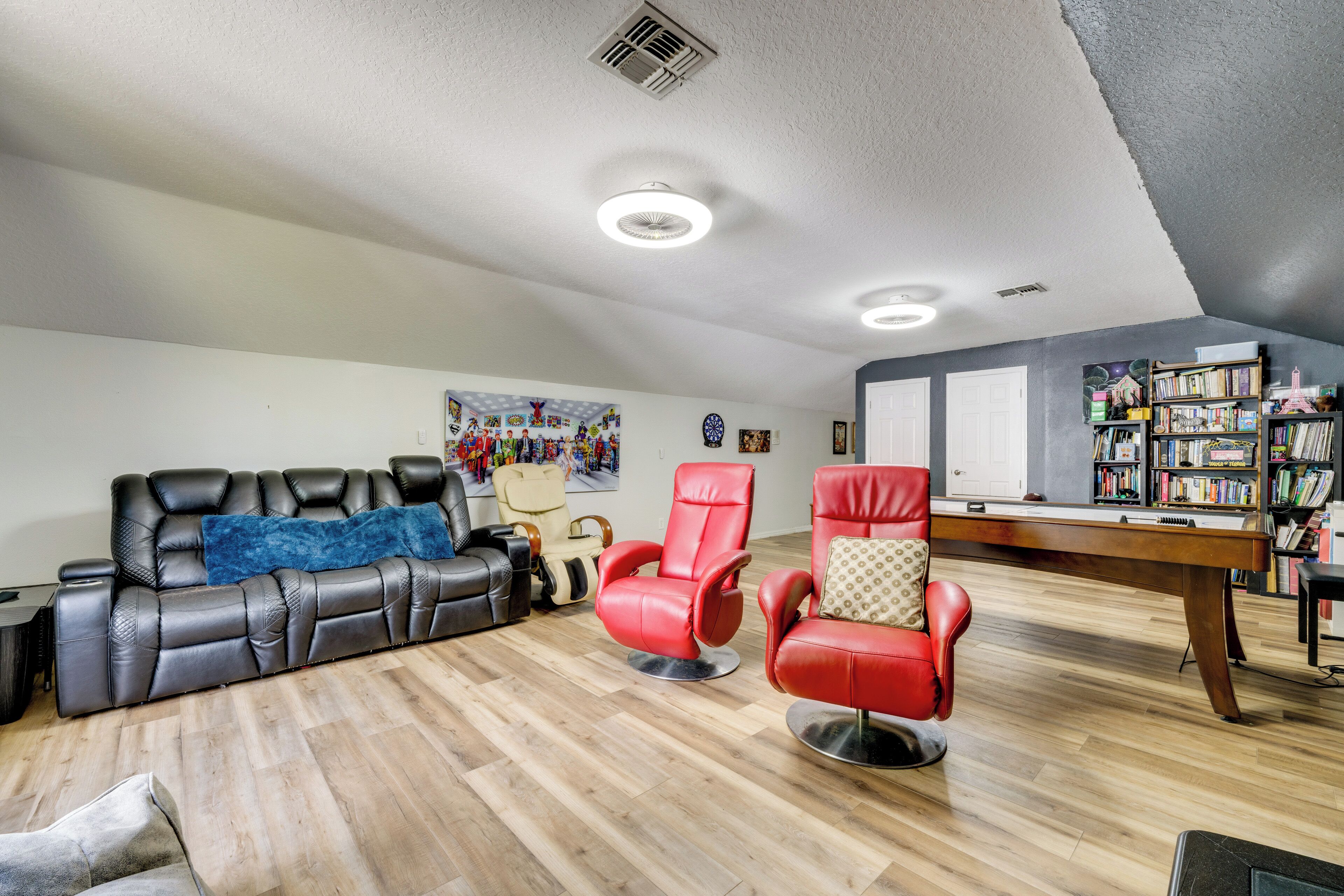 Um quarto com um sofá de couro preto, cadeiras de massagem vermelhas, piso de madeira, uma estante e um alvo de dardos.