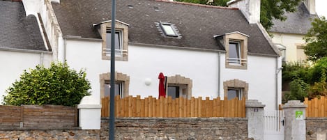 Une maison à deux étages, avec des fondations en pierres, une clôture en bois et une porte rouge.
