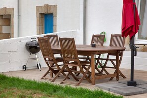 An outdoor wooden table with chairs, a barbecue grill, and a red umbrella.