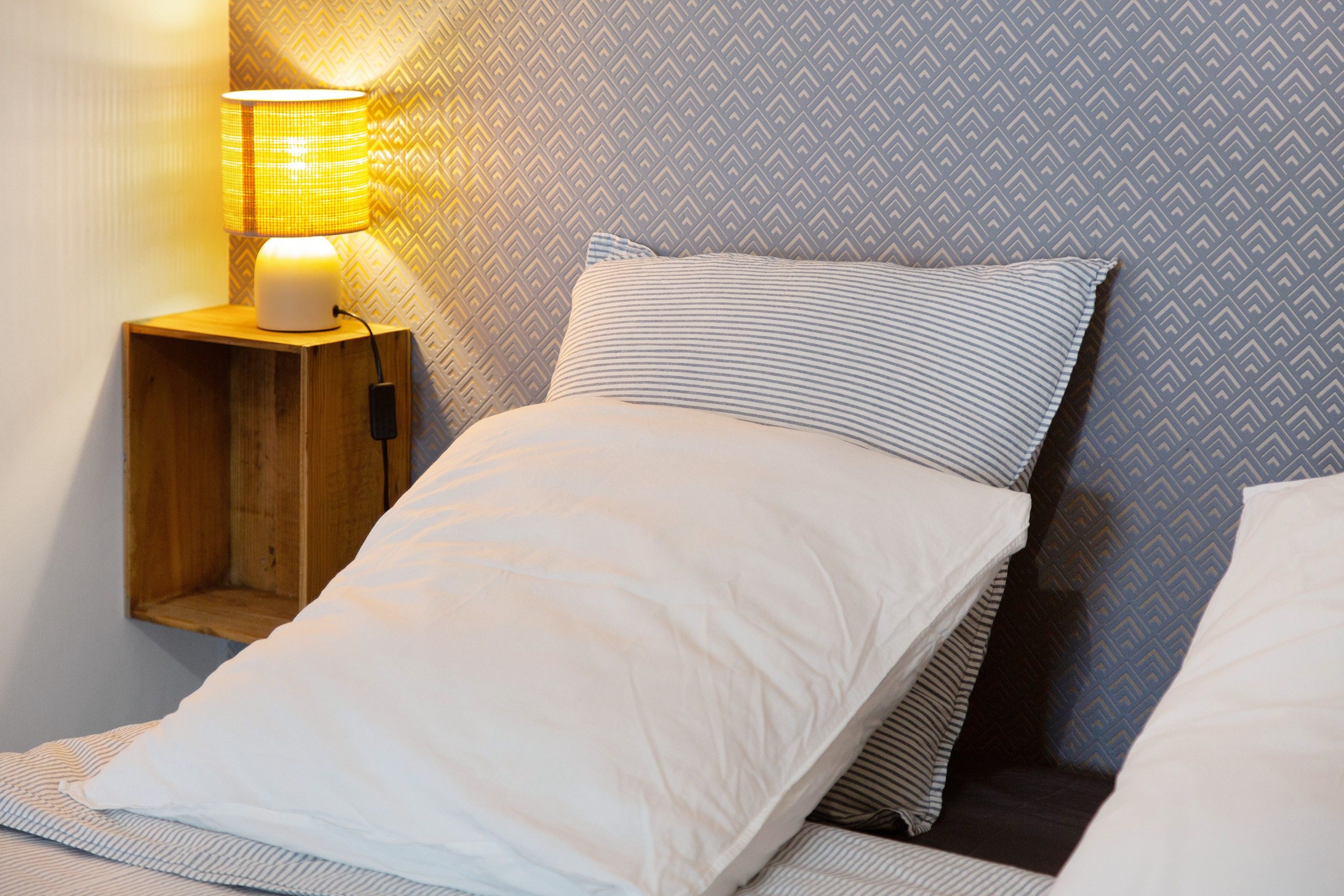 Een bed met wit en gestreept beddengoed, een houten nachtkastje met een lamp, en een kamer met een decoratieve wand.