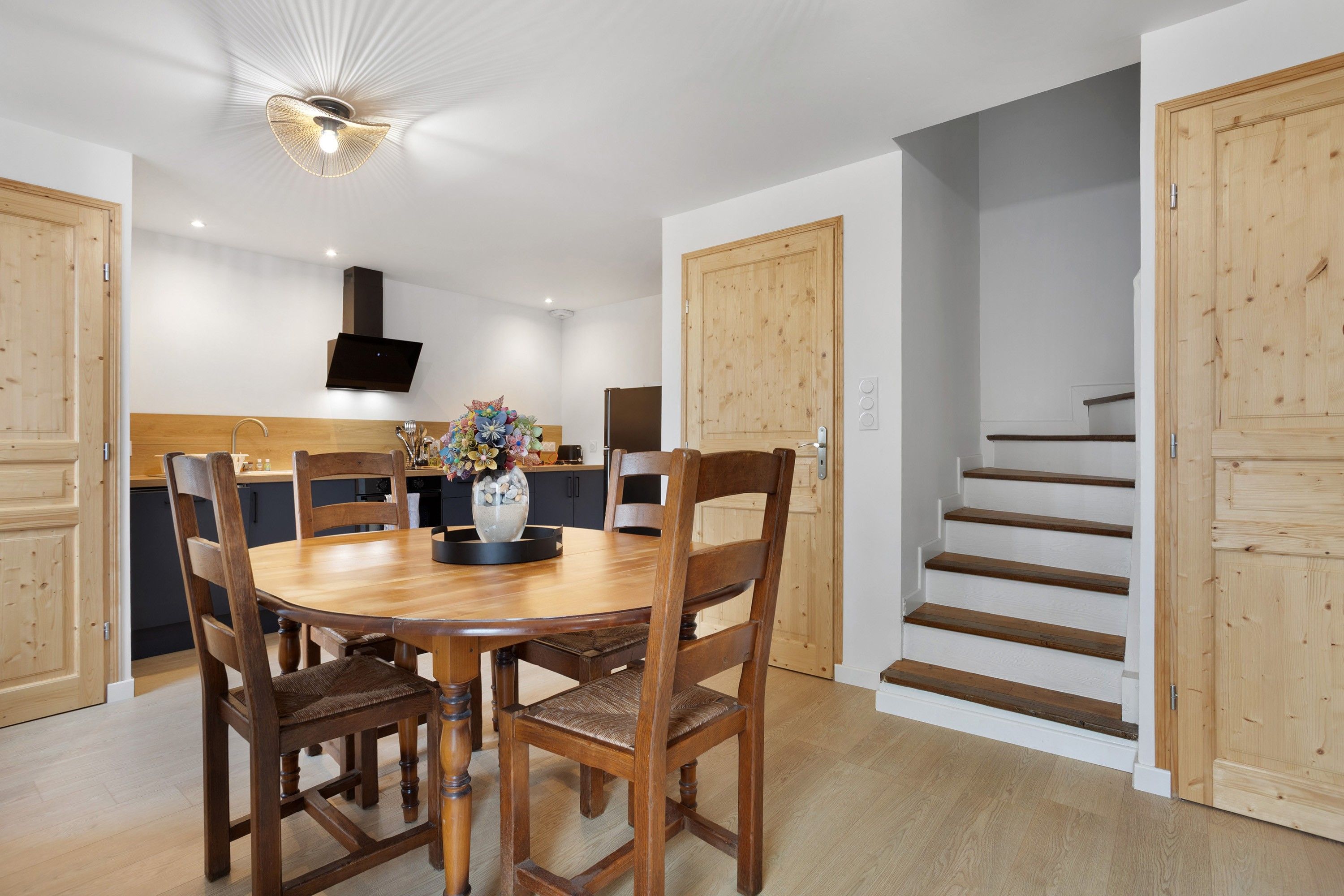 Un coin repas avec une table ronde, des chaises en bois, un vase de fleurs et un luminaire moderne au plafond.