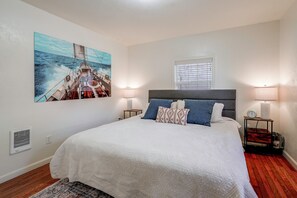 A bedroom with a large bed, two bedside lamps, a window with blinds, a wall-mounted air vent, and a painting of a sailboat. - Walk to Dtwn Corvallis: Home Near OSU & Riverwalk (Corvallis)