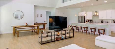 Uma sala ampla com uma mesa de jantar, um sofá, uma TV e uma cozinha ao fundo.