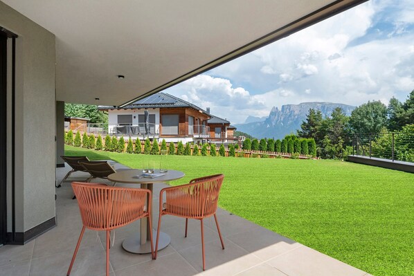 A modern outdoor patio with a dining table and chairs, a view of a mountain range, and a well-maintained lawn.