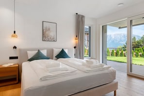 Une chambre moderne avec un grand lit, deux oreillers, une table de chevet avec un livre, et une vue sur un paysage montagneux par une porte-fenêtre coulissante.