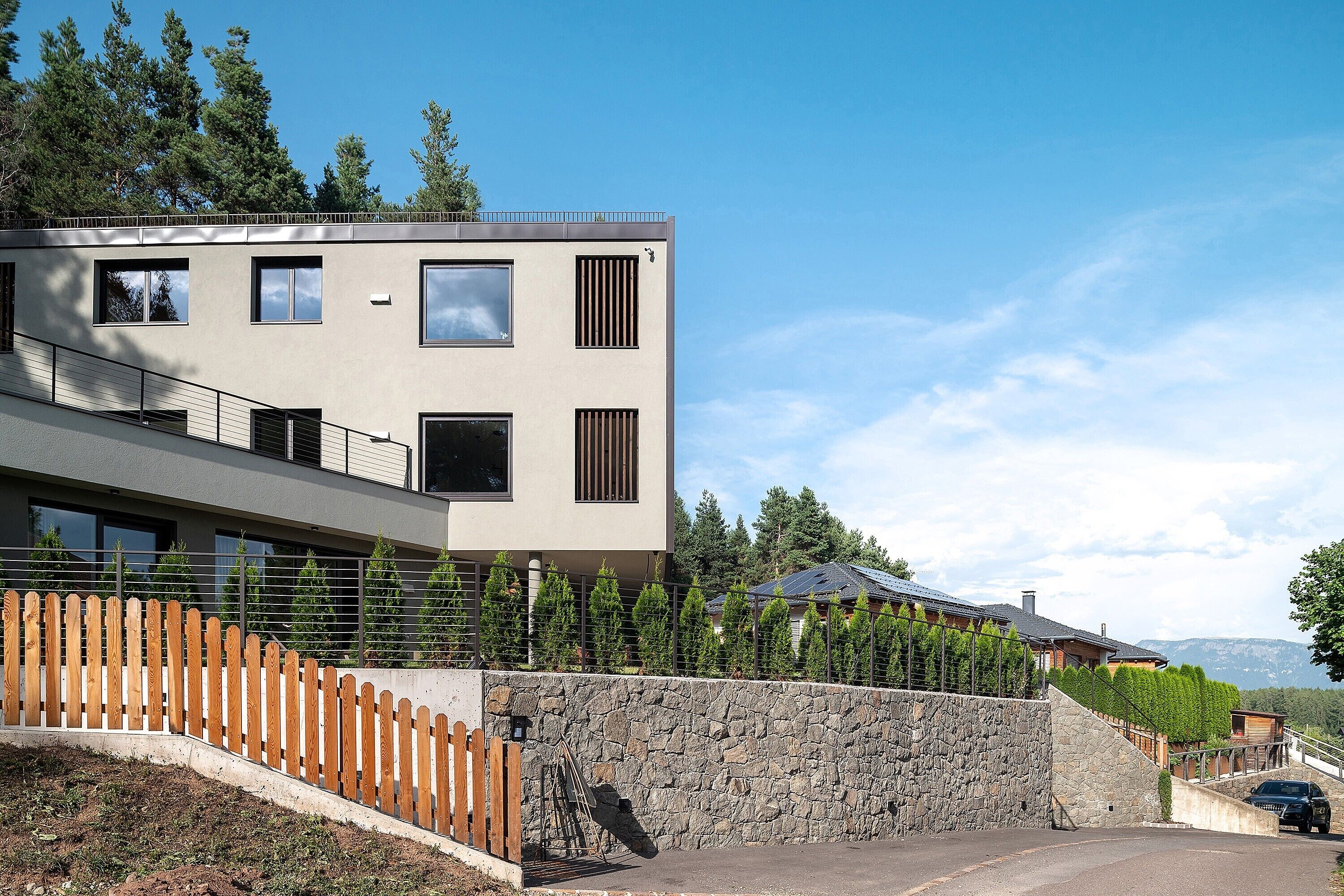 A modern two-story building with a stone retaining wall, a wooden fence, and a view of a mountain range.