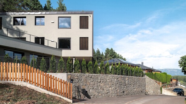 Un edificio moderno de dos pisos, con un muro de contención de piedra, cerca de madera y vistas a una cordillera.