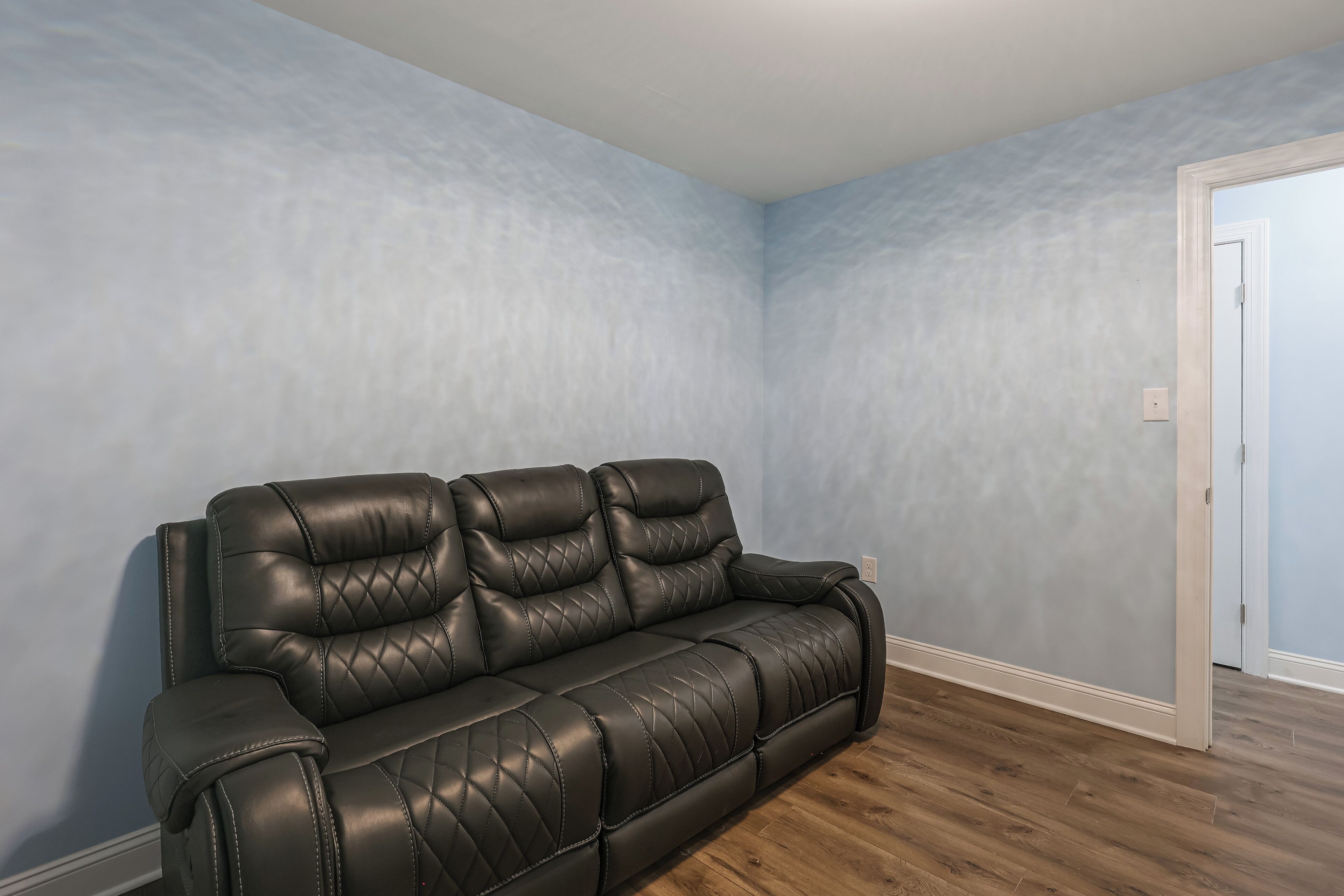 A room with a dark brown leather sofa against light blue walls, a wooden floor, and a white door.