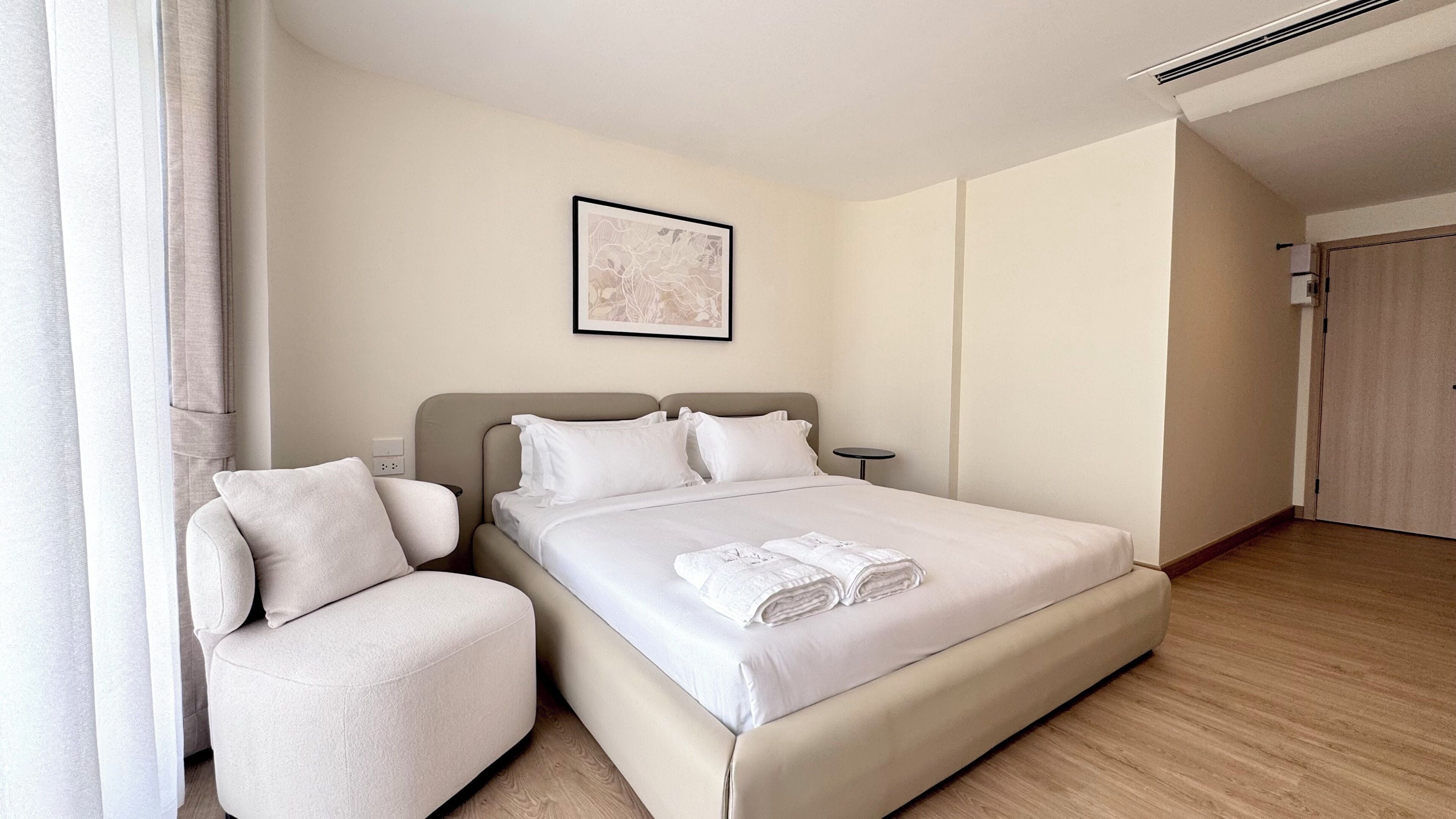 A hotel room with a bed, a bedside table, a chair, a framed picture on the wall, and a door.