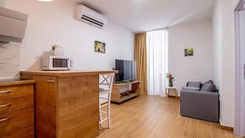 Un espacio habitable compacto con kitchenette, un televisor de pantalla plana, un sofá gris y una mesita auxiliar pequeña.