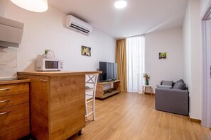 Un espacio habitable compacto con kitchenette, un televisor de pantalla plana, un sofá gris y una mesita auxiliar pequeña.