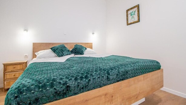 A single bed with a wooden frame and a green textured bedspread, flanked by two nightstands and a wall-mounted light fixture.