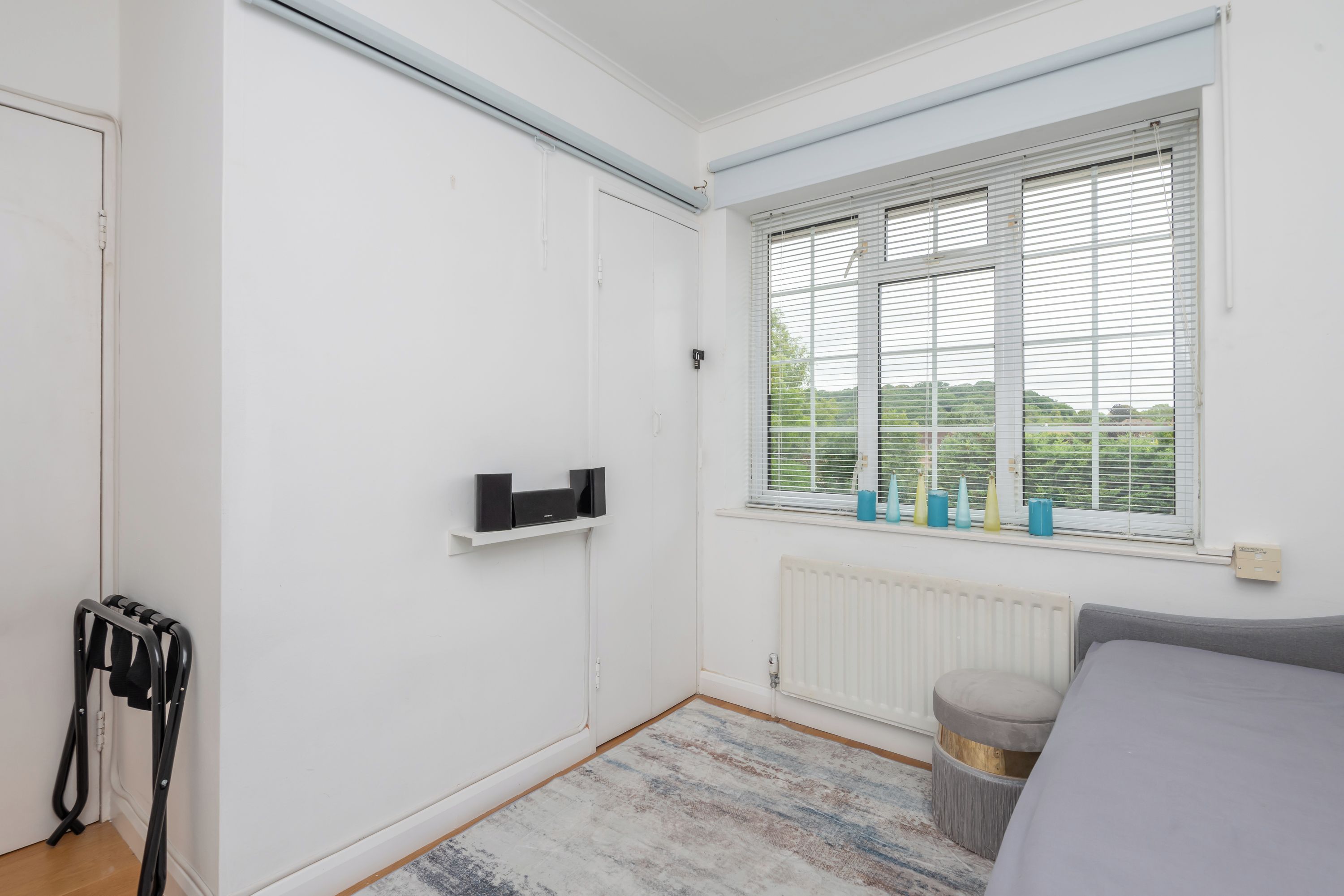 A bedroom with a white wardrobe, a radiator, a window with blinds, and a grey sofa.