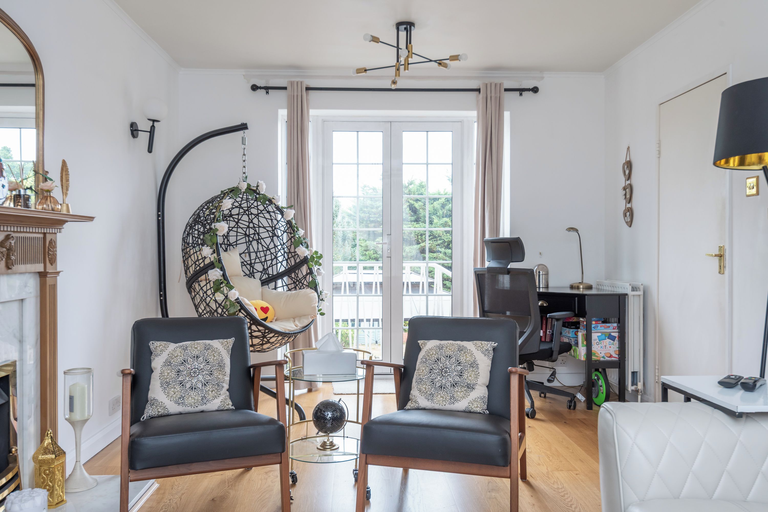 A living room with two black leather chairs, a glass coffee table, a hanging chair with white flowers, a fireplace, and a view of the outdoors through a large window.