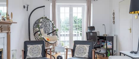 Una sala de estar con dos sillones de cuero negro, una mesa de centro de vidrio, una silla colgante adornada con flores blancas, una chimenea y vistas al exterior a través de un amplio ventanal.