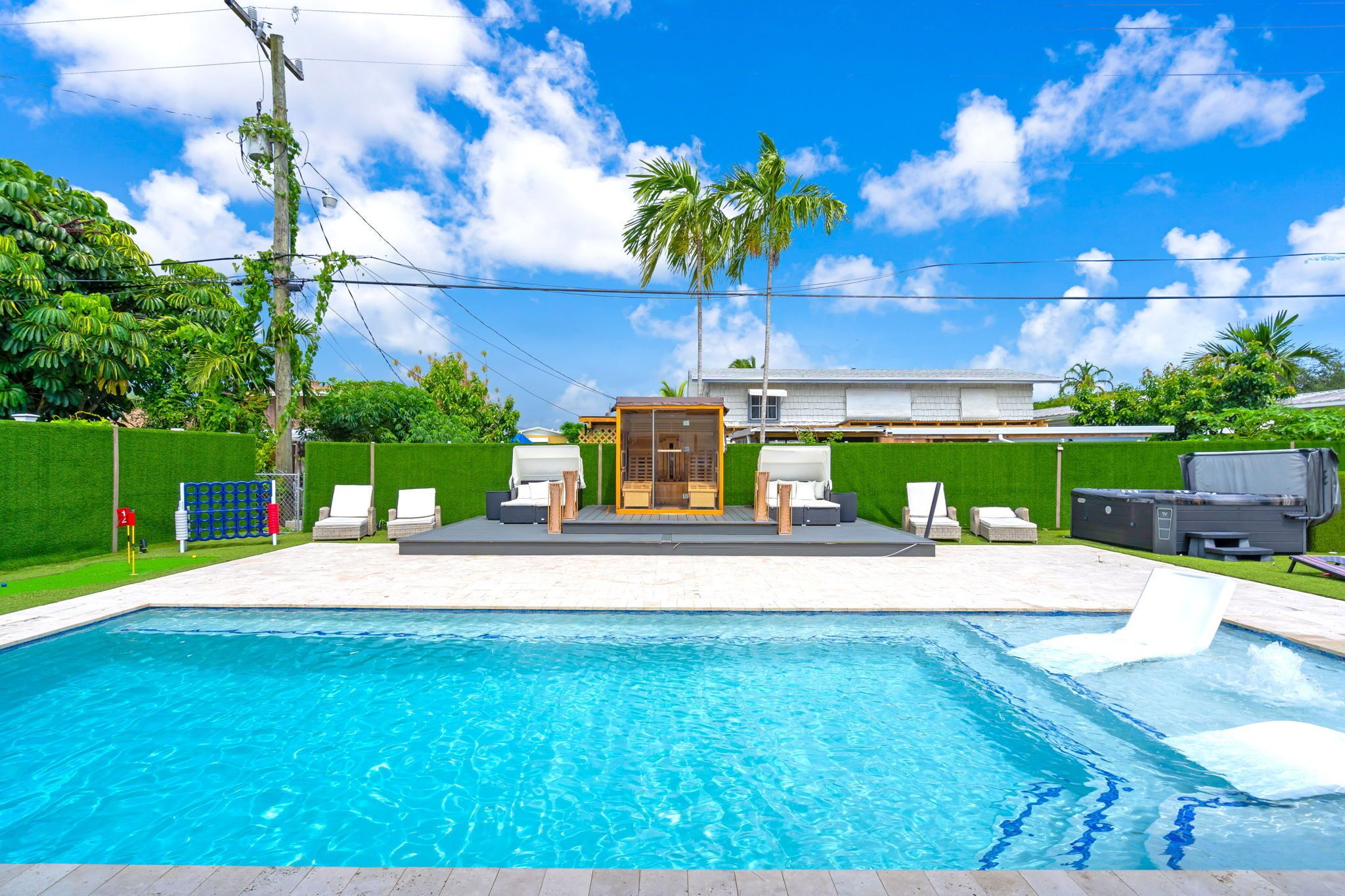 A swimming pool with a sun deck area, surrounded by a green hedge and palm trees.