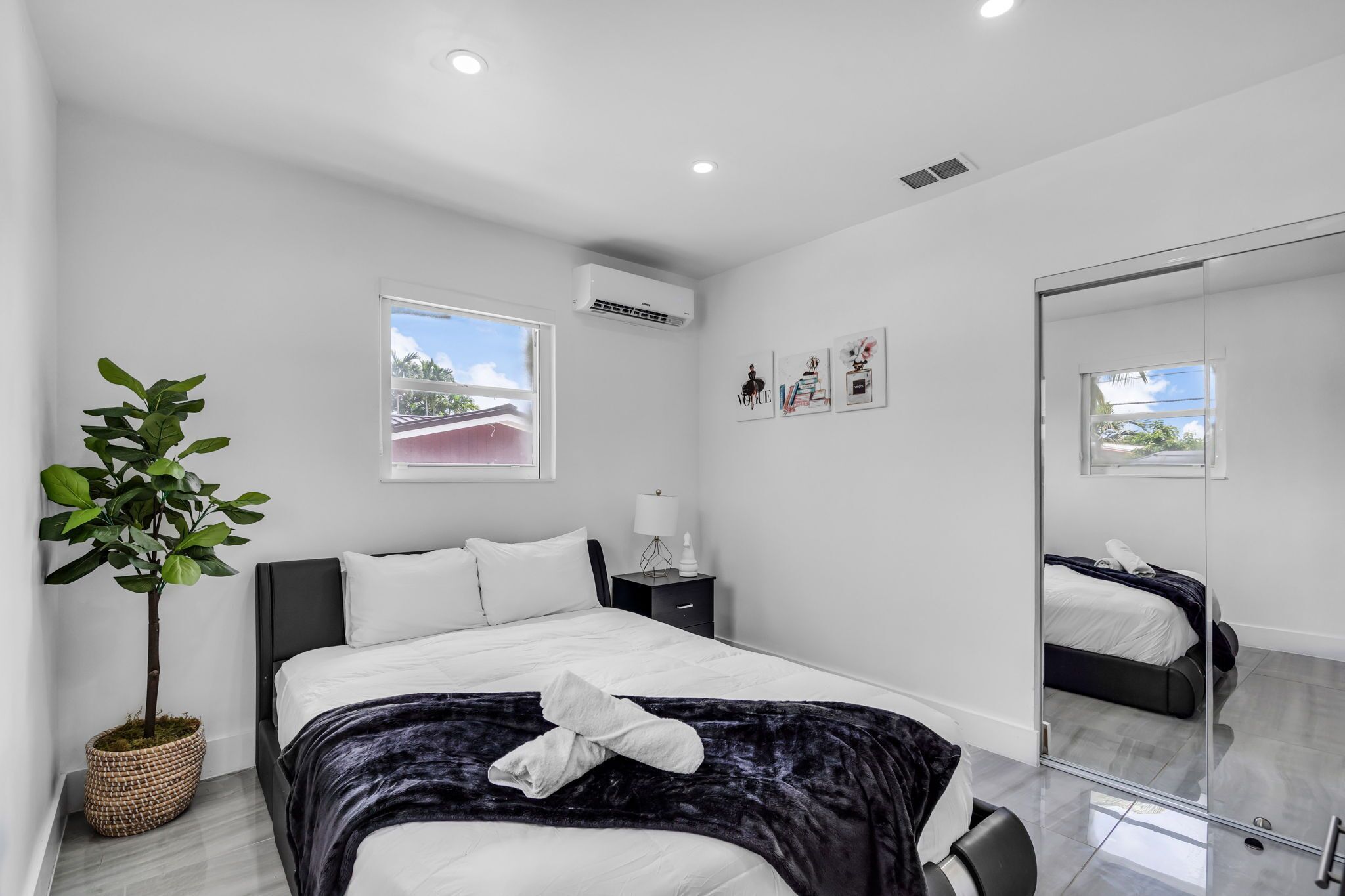 Une chambre moderne comprenant un lit, une table de chevet, une lampe, un appareil de climatisation, une fenêtre et un grand miroir.