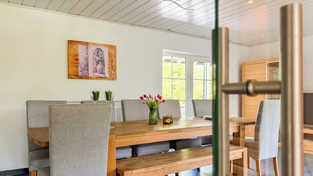 A dining area with a wooden table, grey upholstered chairs, and a bench. There is a painting on the wall and a window with a view of greenery. - Spacious, detached villa for 12 people near Winterberg (Grönebach)