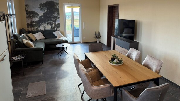 A modern living room with a sofa, a dining table, and a TV. - Newly Built Farmhouse-Style Vacation Rental Near the Borders of Luxembourg and Belgium (Üttfeld)