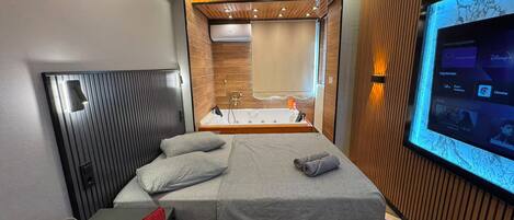 A modern hotel room with a bed, a TV, a bathroom, and a seating area.
