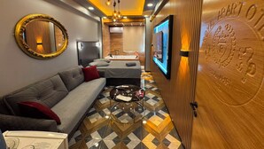 A hotel room with a sofa, a coffee table, a mirror, a TV mounted on the wall, and a wooden door with a plaque.