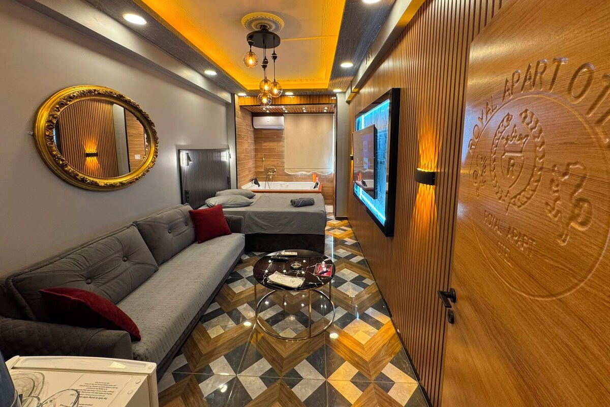 A hotel room with a sofa, a coffee table, a mirror, a TV mounted on the wall, and a wooden door with a plaque.