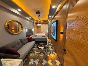 A hotel room with a sofa, a coffee table, a mirror, a TV mounted on the wall, and a wooden door with a plaque.