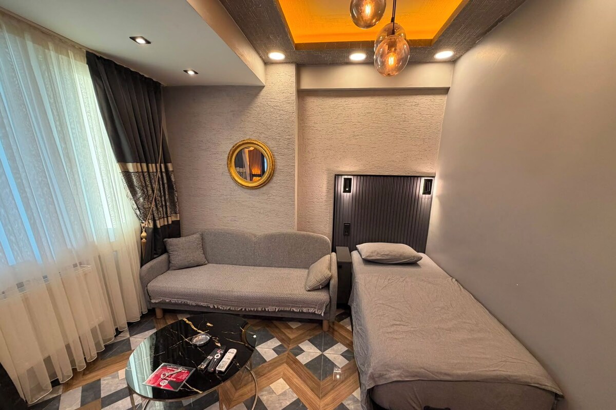 A compact hotel room with a sofa, a single bed, a small table with remote controls, and a patterned floor.