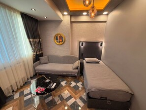 A compact hotel room with a sofa, a single bed, a small table with remote controls, and a patterned floor.