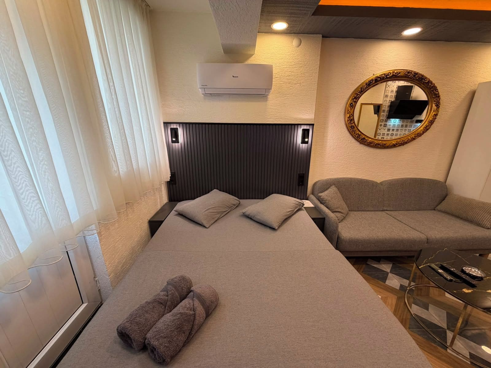 A hotel room with a bed, a sofa, a glass coffee table, and an air conditioning unit.