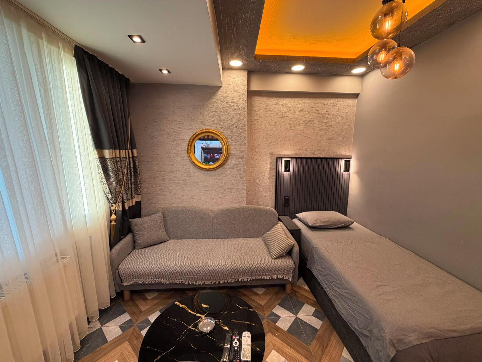 Une pièce de taille modeste, comprenant un canapé, un lit, un miroir rond et une table basse avec une télécommande.