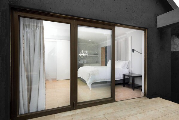 Un dormitorio moderno con un amplio ventanal corredera que da a un baño, una cama con ropa blanca y una mesita de noche con lámpara.