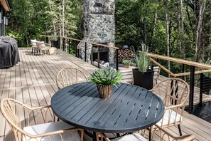 Una terraza de madera con mobiliario exterior, una chimenea de piedra y vistas del bosque circundante.