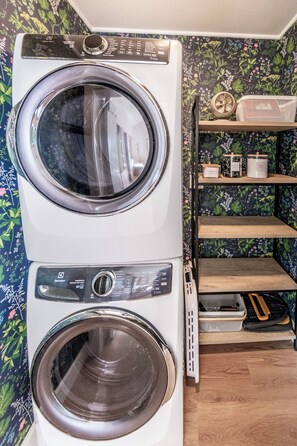 A stacked washer and dryer unit with a control panel, a shelf with storage containers, and a floral wallpaper background. - The Aspens at Meadow Creek (Anchorage)