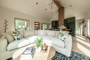 Una sala moderna con un sofá seccional curvo, una mesa de centro con libros y una planta, y una cocina al fondo.