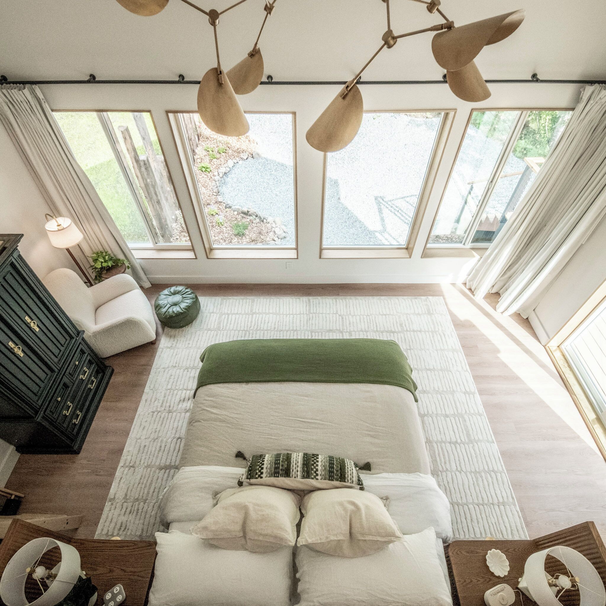 A bedroom with a large bed, a dresser, a lamp, and a view of the outdoors through large windows.