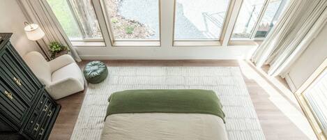 Un dormitorio con una cama grande, un aparador, una lámpara y vistas al exterior a través de amplios ventanales.