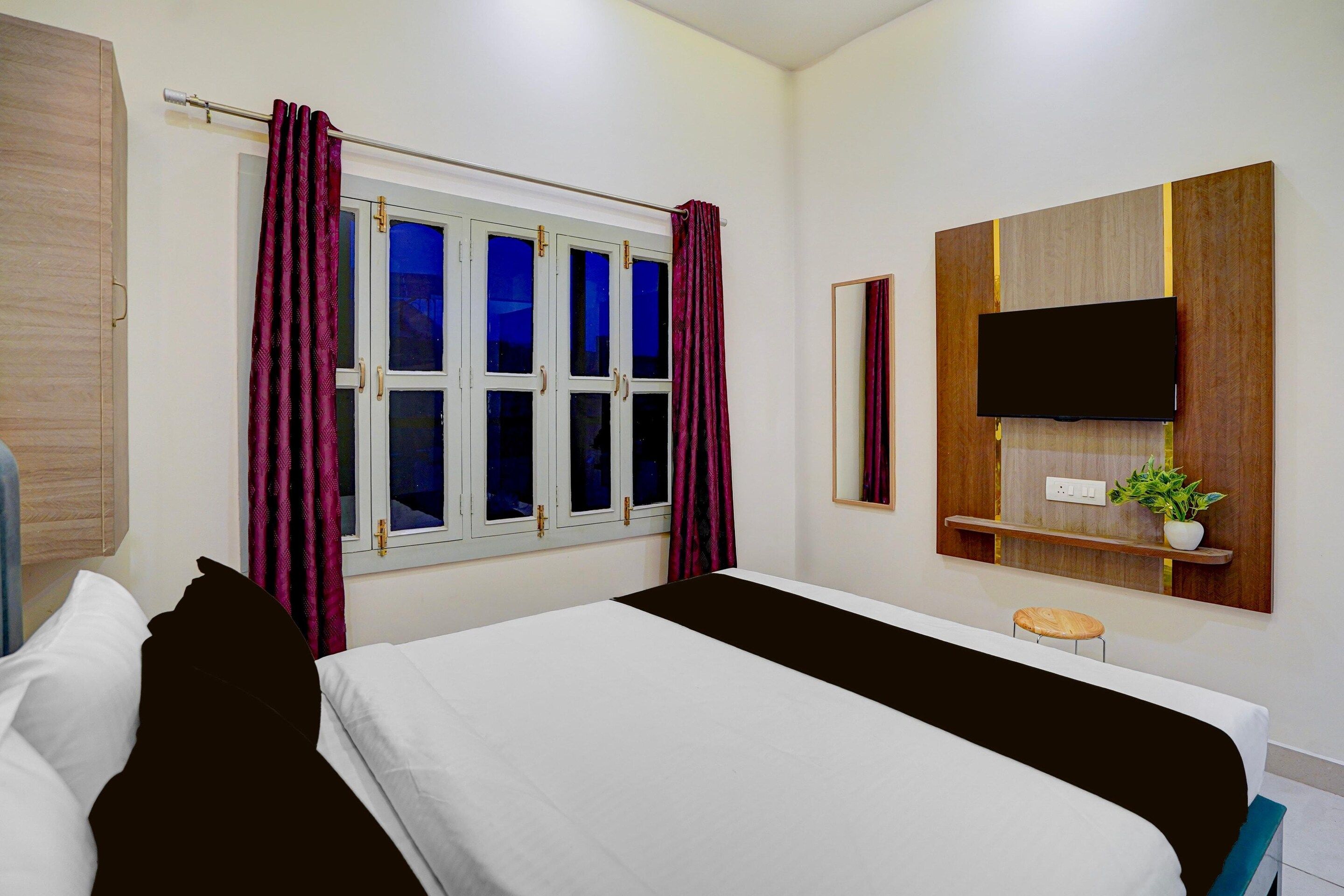 Una habitación de hotel con una cama, un ventanal con cortinas, un televisor montado en la pared, un taburete pequeño y una planta en un estante.