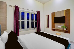 Een hotelkamer met een bed, een raam met gordijnen, een aan de muur bevestigde televisie, een klein krukje en een plant op een plank.