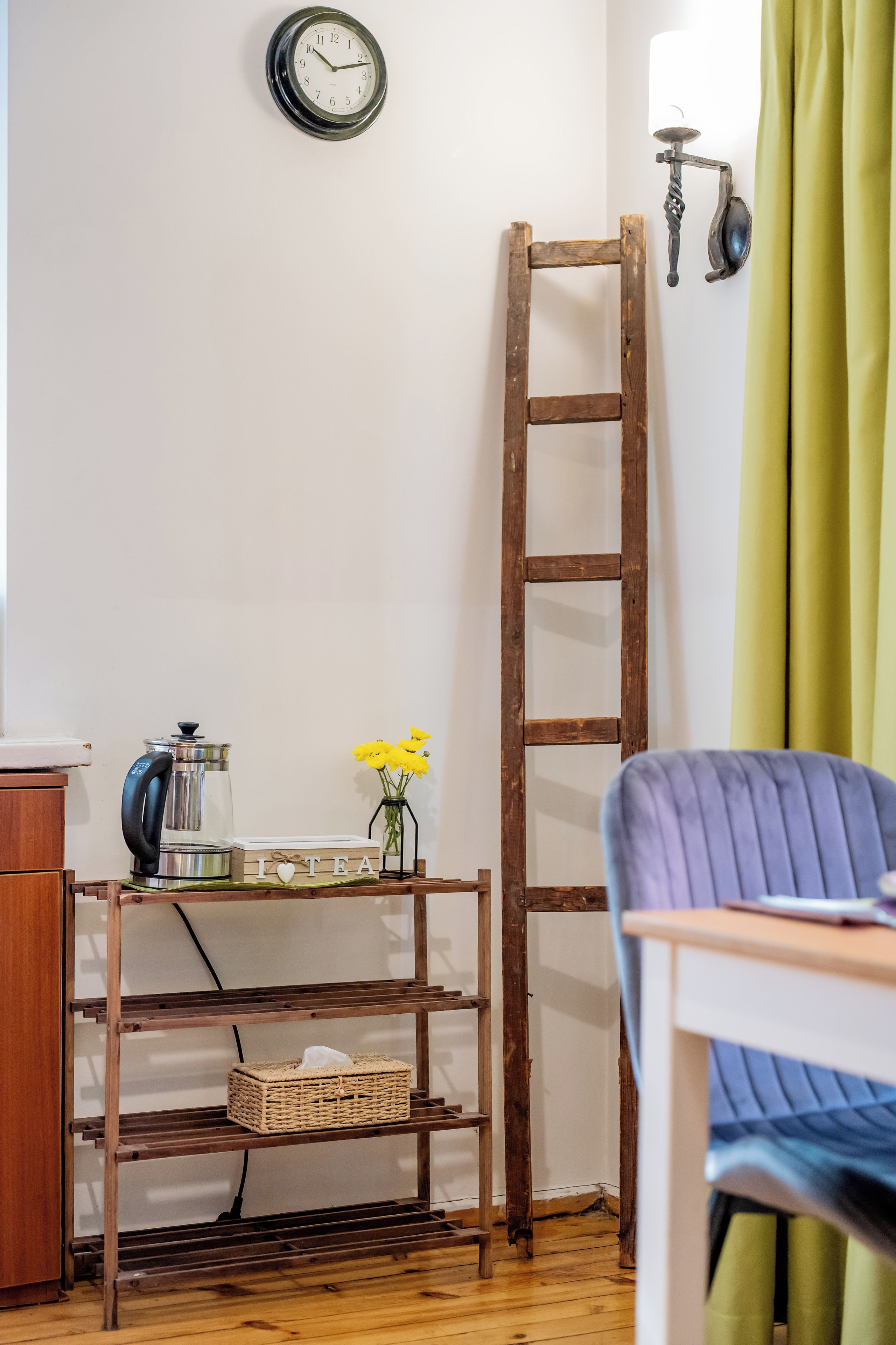 A room with a wooden ladder, a shelf with a kettle and flowers, a clock on the wall, and a chair.