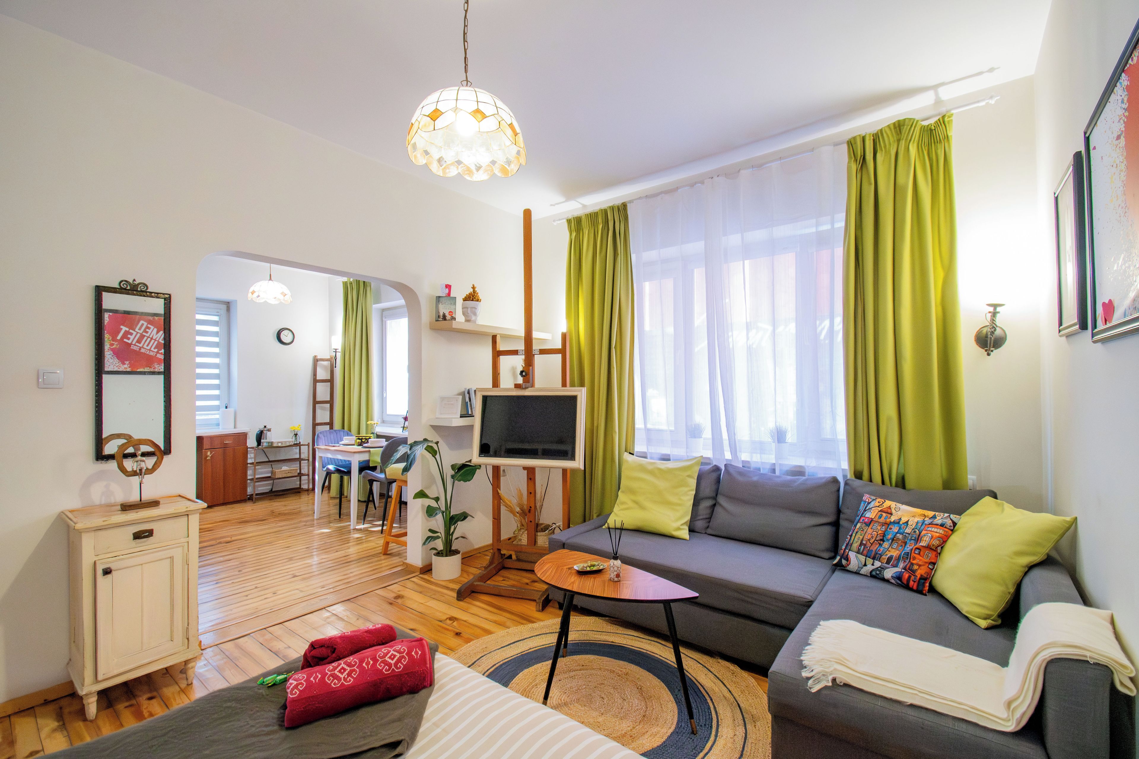 A modern living room with a sofa, a small table, a TV, and a dining area in the background.