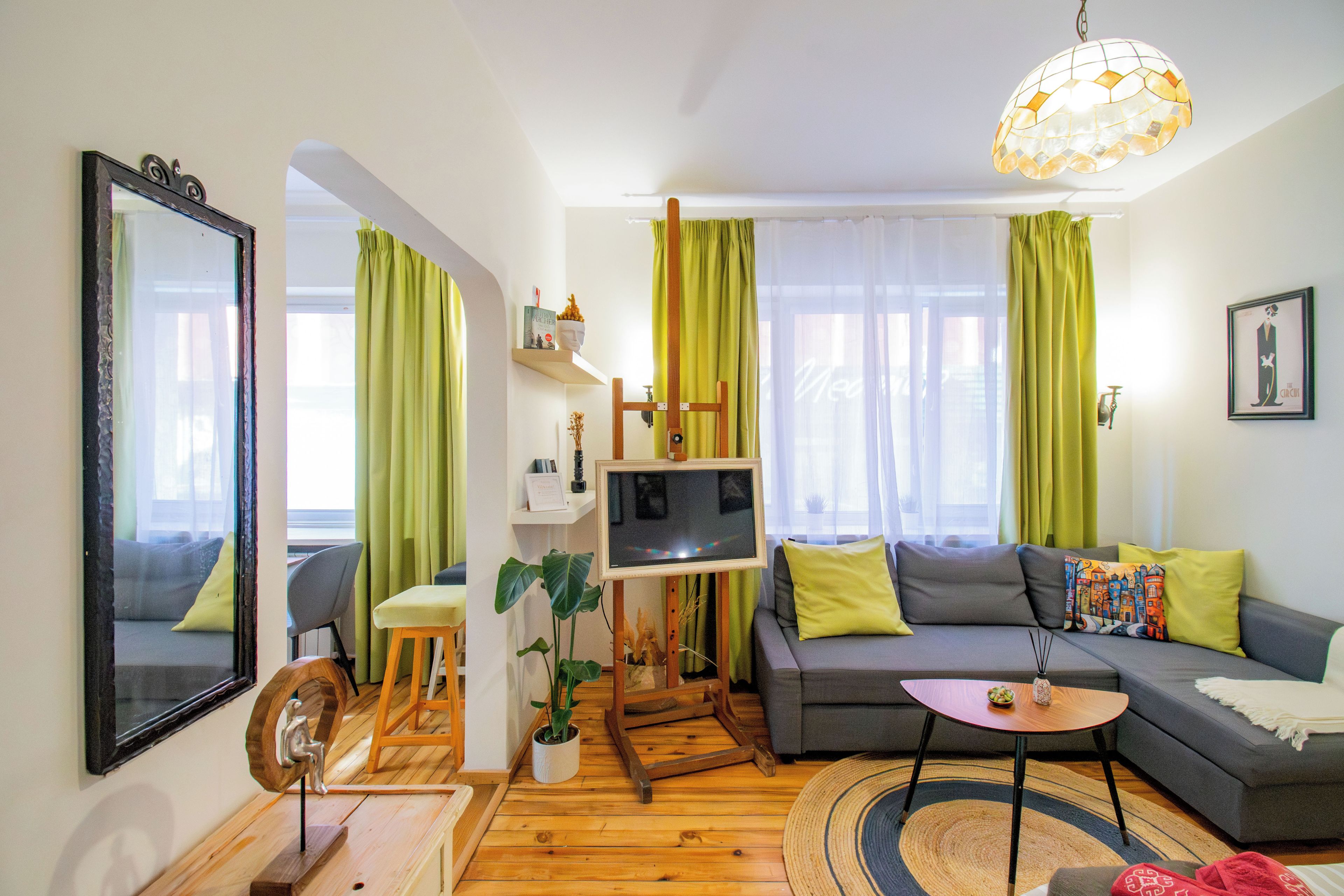 A modern living room with a grey sofa, a small wooden table, a TV on a stand, and a large mirror on the wall.