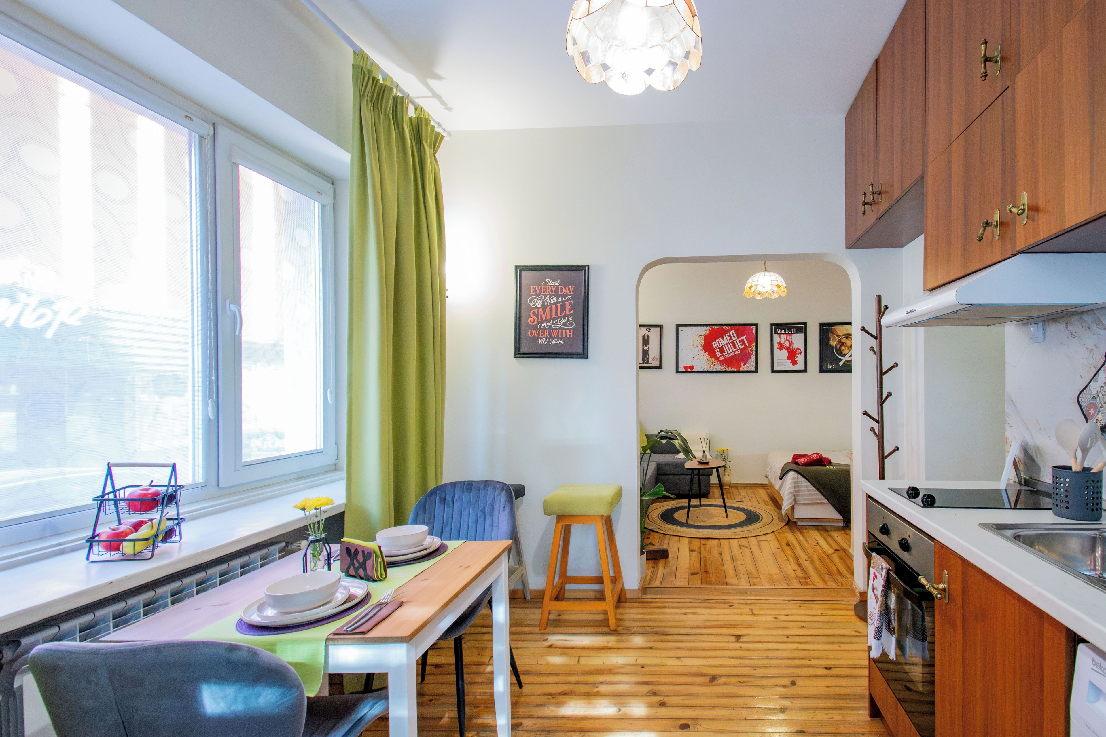 A compact kitchen with wooden flooring, a dining area with a table set for two, and a view of the living room through an open doorway.