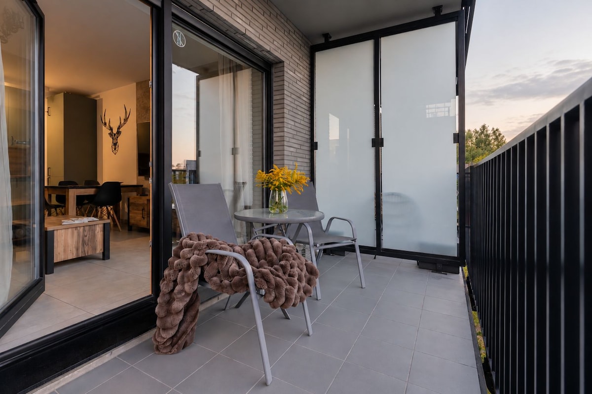 A modern balcony with a glass door leading to an interior dining area, a small round table with a vase of yellow flowers, and a chair with a textured blanket.