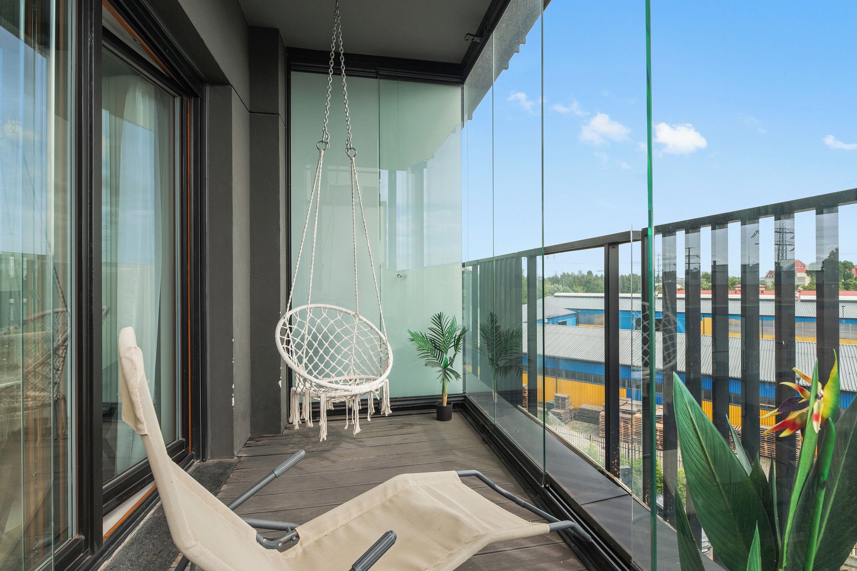 A balcony with a hanging chair, a potted plant, and a view of a blue building.