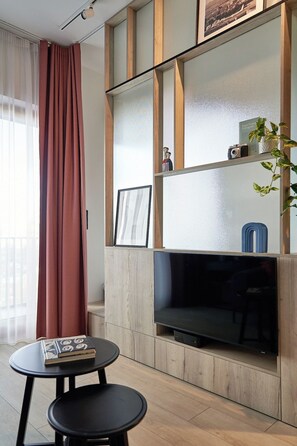 A modern living room with a flat-screen TV, a small table with a book, and a window with curtains. - Riverside Gdańsk Apartment by Lion Apartments (Gdańsk)